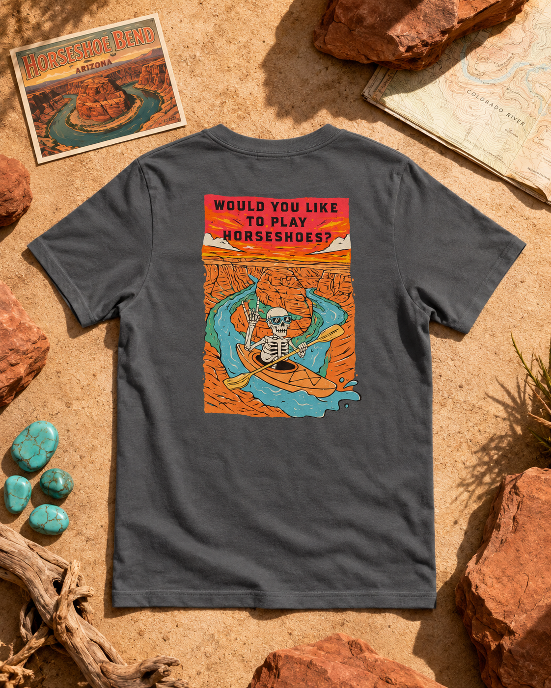 Solid Asphalt Blend Do You Wanna Play Horseshoes? T-shirt featuring the skeleton kayaker Horseshoe Bend back graphic, flat lay on red rock with hiking gear
