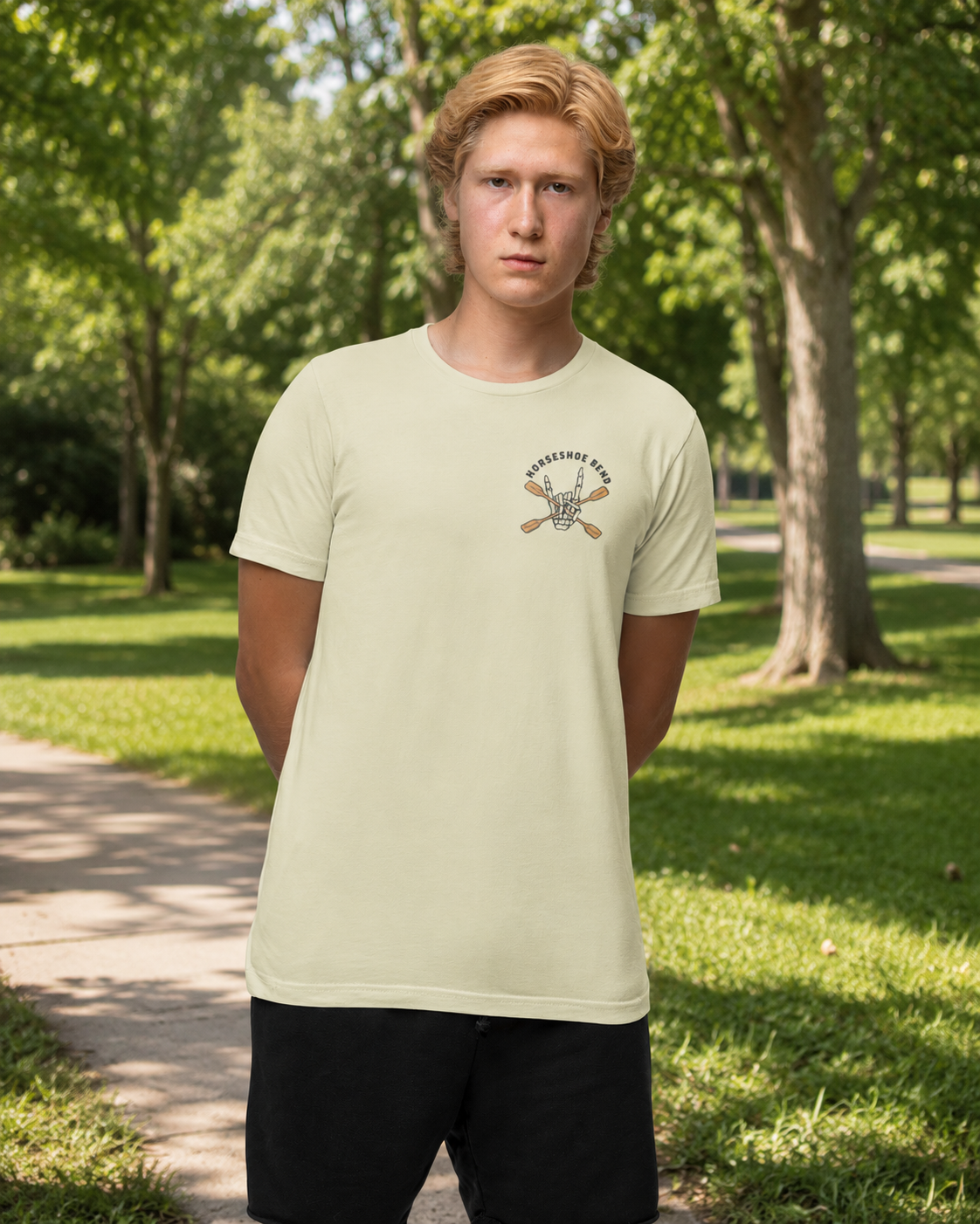 Man wearing a French vanilla Do You Wanna Play Horseshoes? T-shirt with small skeleton kayaker chest graphic in a sunny park setting