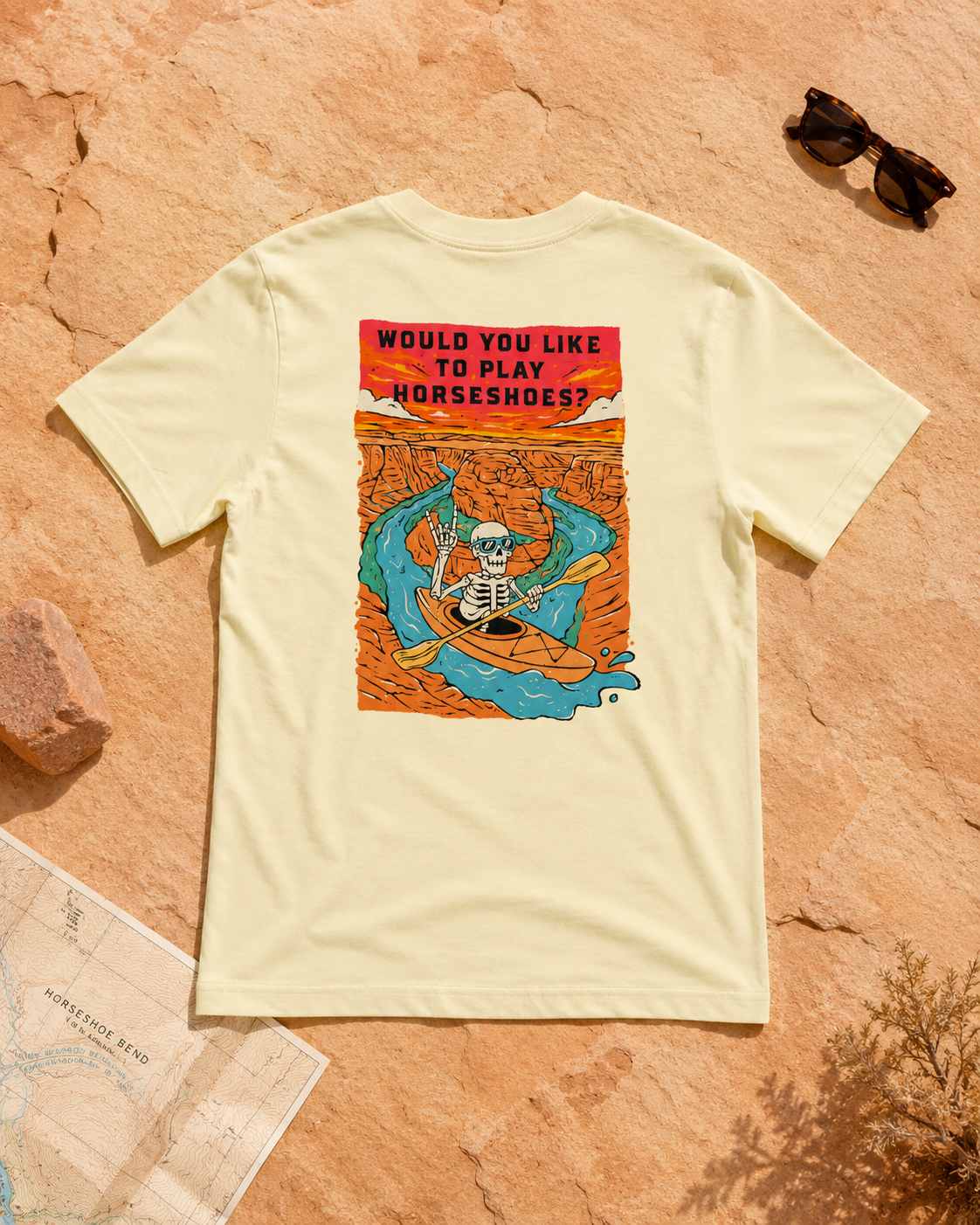 French Vanilla Do You Wanna Play Horseshoes? T-shirt featuring the skeleton kayaker Horseshoe Bend back graphic, flat lay on desert rock