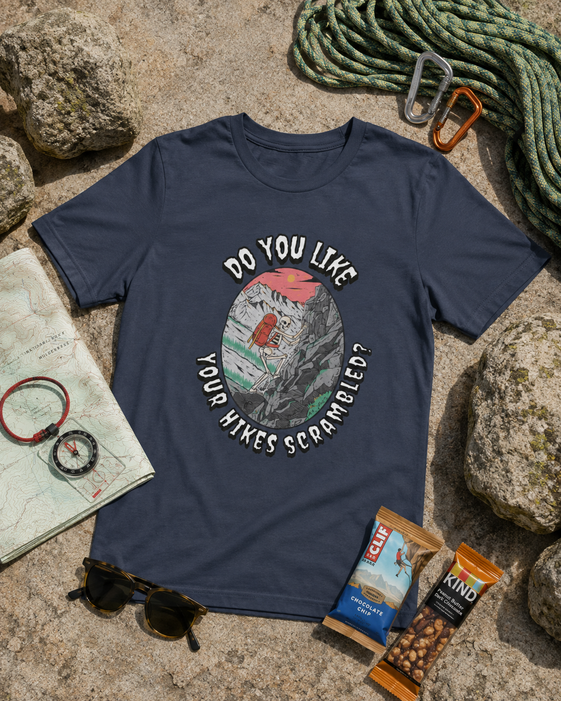 Heather Navy Do You Like Your Hikes Scrambled? T-shirt featuring a skeleton scrambling over rocks, flat lay with outdoor hiking gear