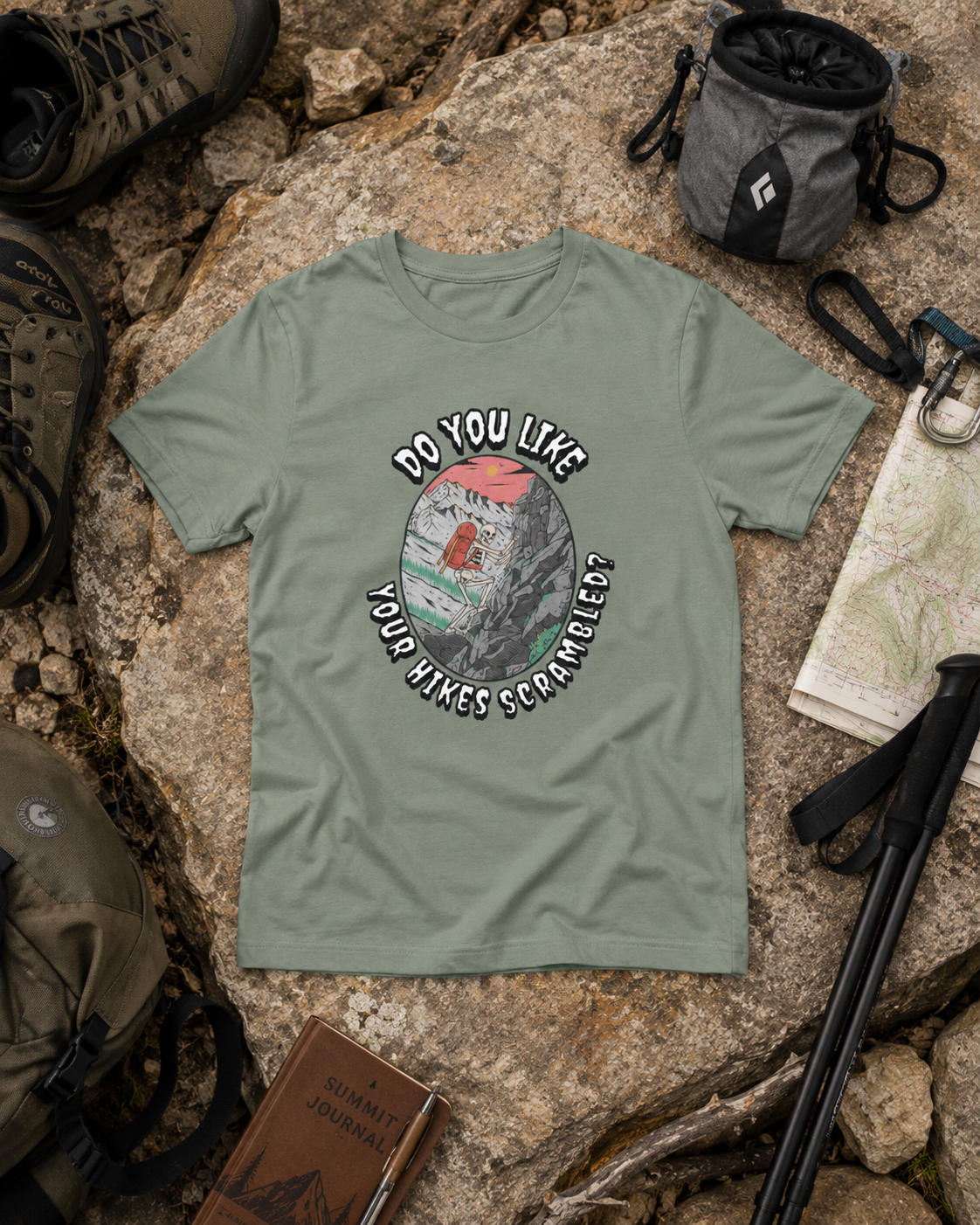 Heather Sage Do You Like Your Hikes Scrambled? T-shirt featuring a skeleton scrambling over rocks, flat lay with hiking and climbing gear